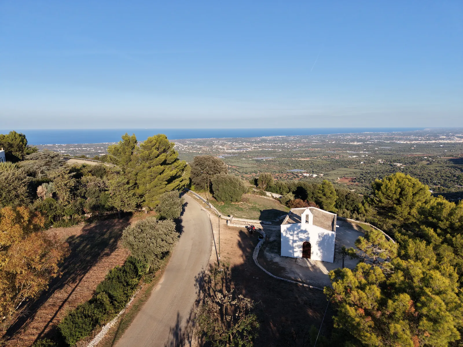 La chiesa bianca vista dall’alto, con il piazzale rotondo e la strada che scende, i pini intorno e la piana fino al mare.