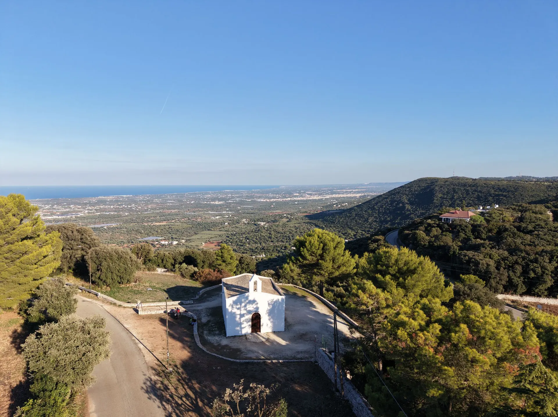 La chiesa bianca sul ciglio, con il piazzale rotondo, la strada asfaltata, il colle boscoso a destra e la piana fino al mare.