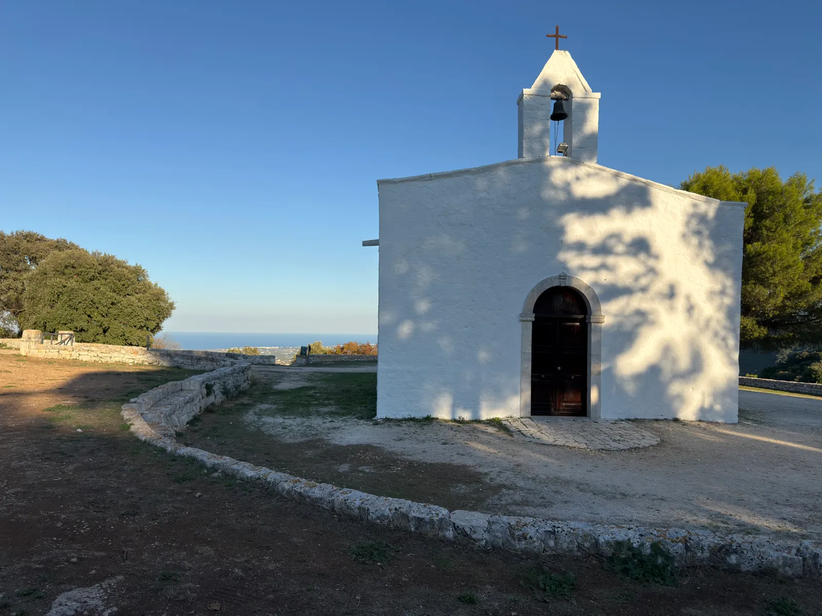La facciata bianca della chiesa con il campanile a vela e la porta ad arco, sul piazzale rotondo, con il mare in lontananza.