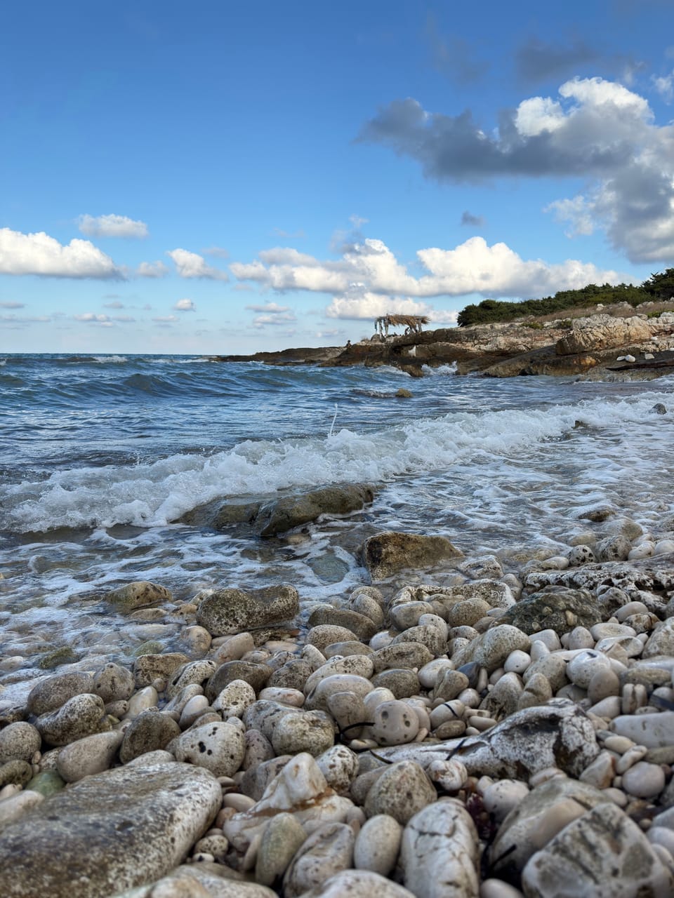 Una spiaggia di ciottoli bianchi con le onde che si rompono sui sassi e una scogliera bassa sullo sfondo.